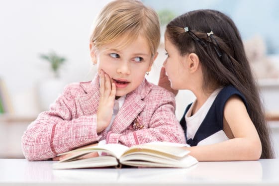Post pic for Blog Writing post Portrait of two little cute girls. Girls sitting at table with book and whispering secrets.